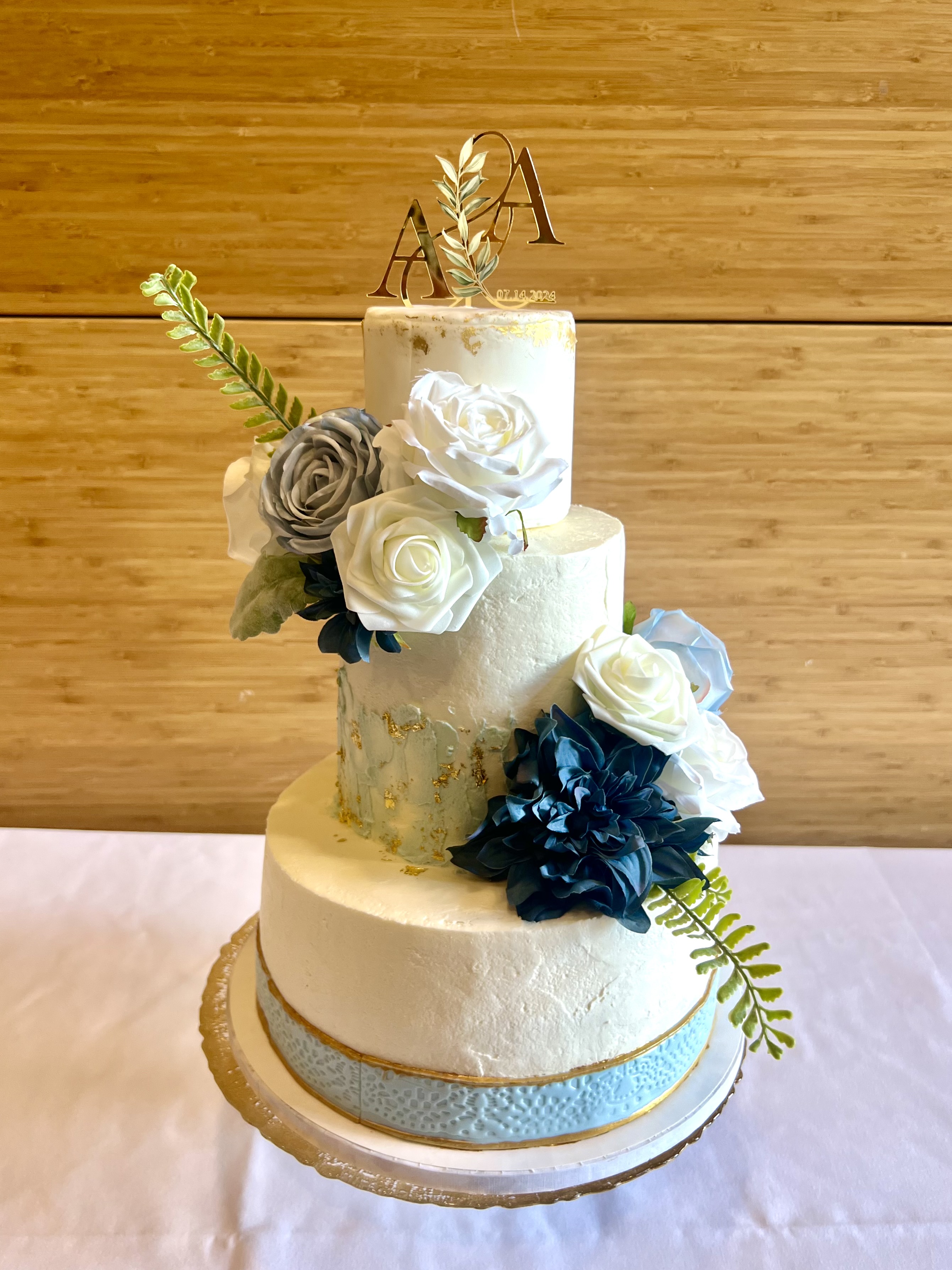 Three-tier wedding cake with gold leaf, blue and white roses, and A&A monogram topper