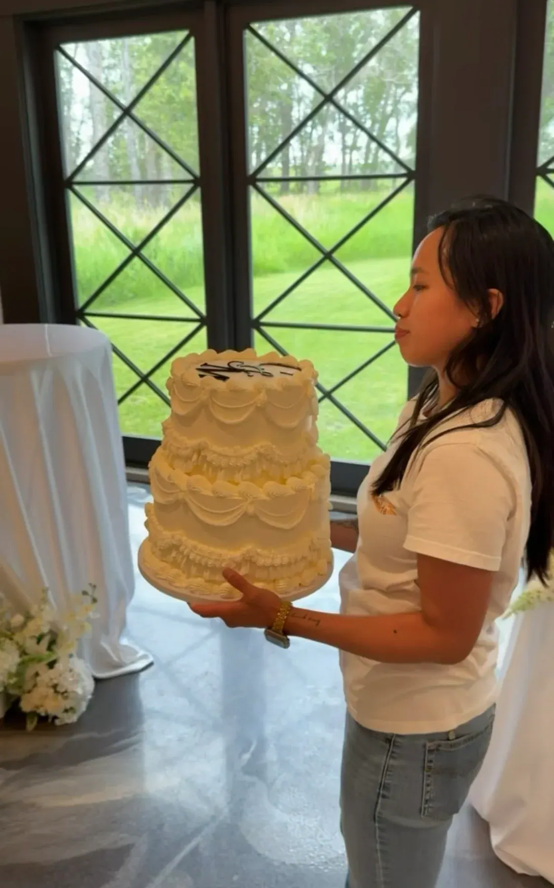 Nicole carrying a vintage-piped ivory two-tier wedding cake through a venue with tall windows