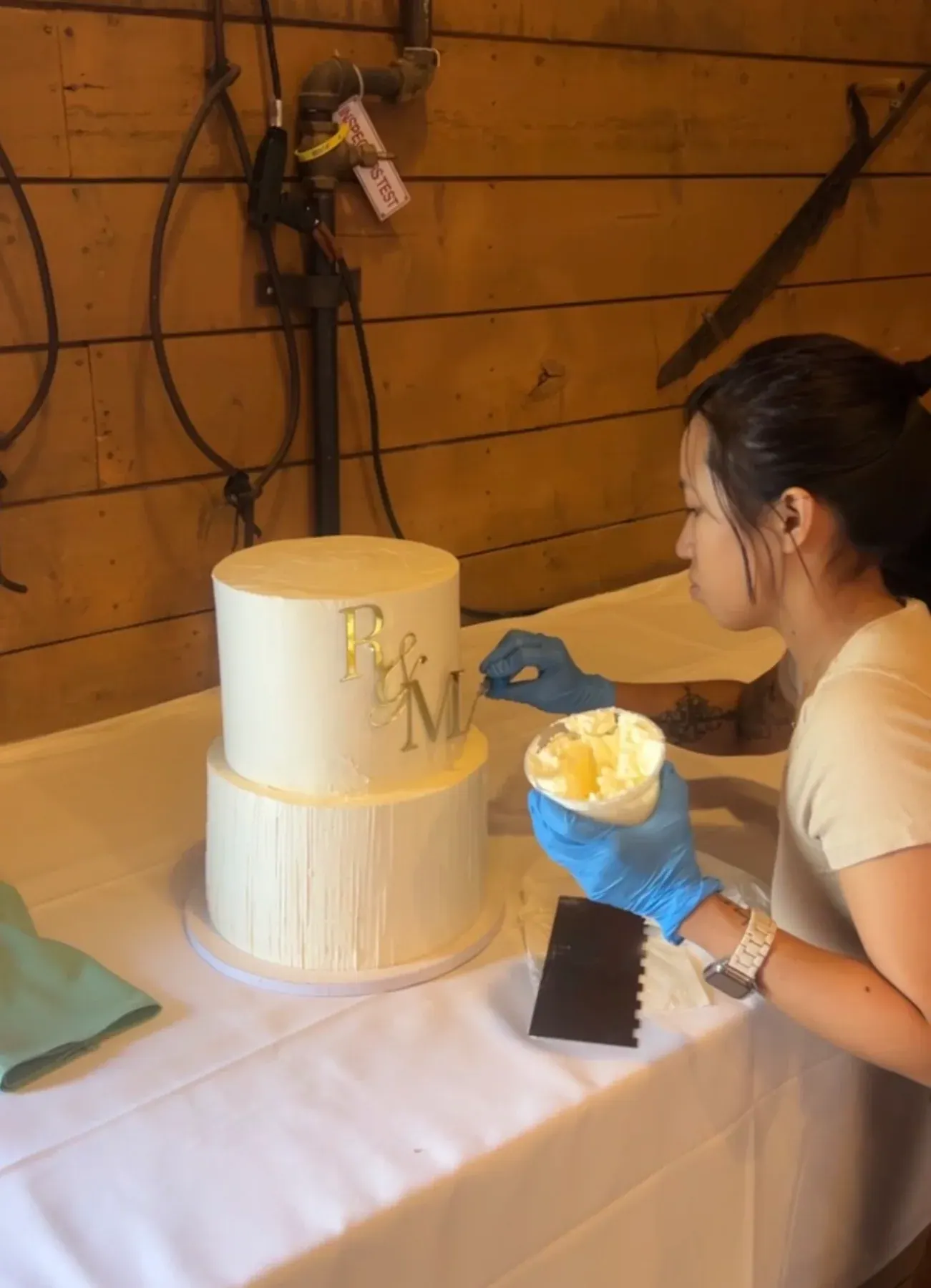 Nicole in gloves adding final buttercream touches to a white two-tier wedding cake with a gold R and M monogram