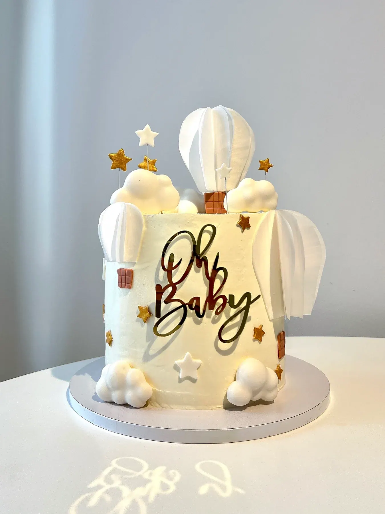 White baby shower cake reading Oh Baby, with a hot-air balloon, clouds and gold stars