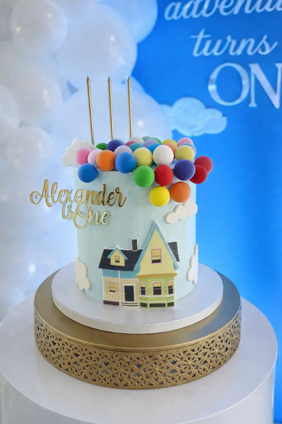 Up-inspired first birthday cake with a little painted house, clouds and a crown of colourful balloons on a gold stand