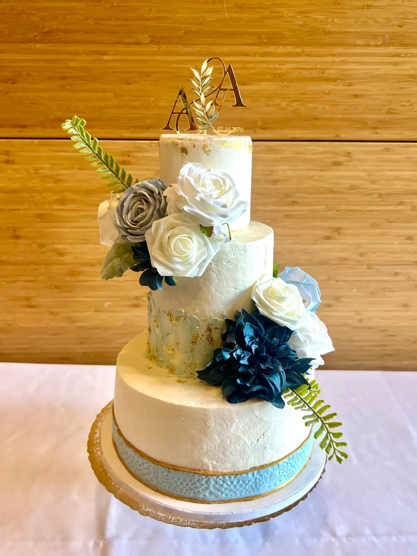 Three-tier ivory wedding cake with blue lace band, gold leaf and blue and white roses