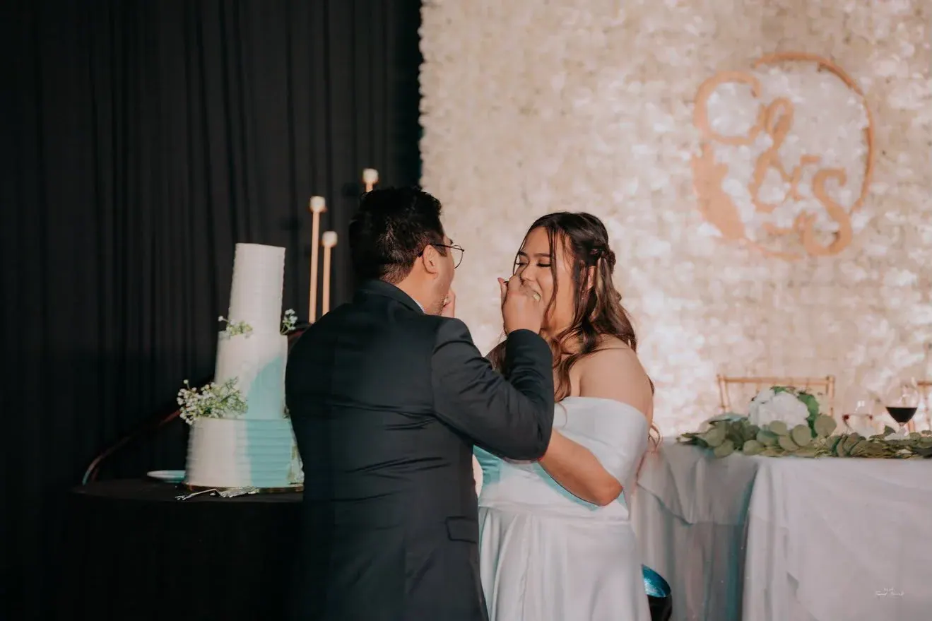 Newlyweds sharing a bite beside their tall white wedding cake in front of a flower wall