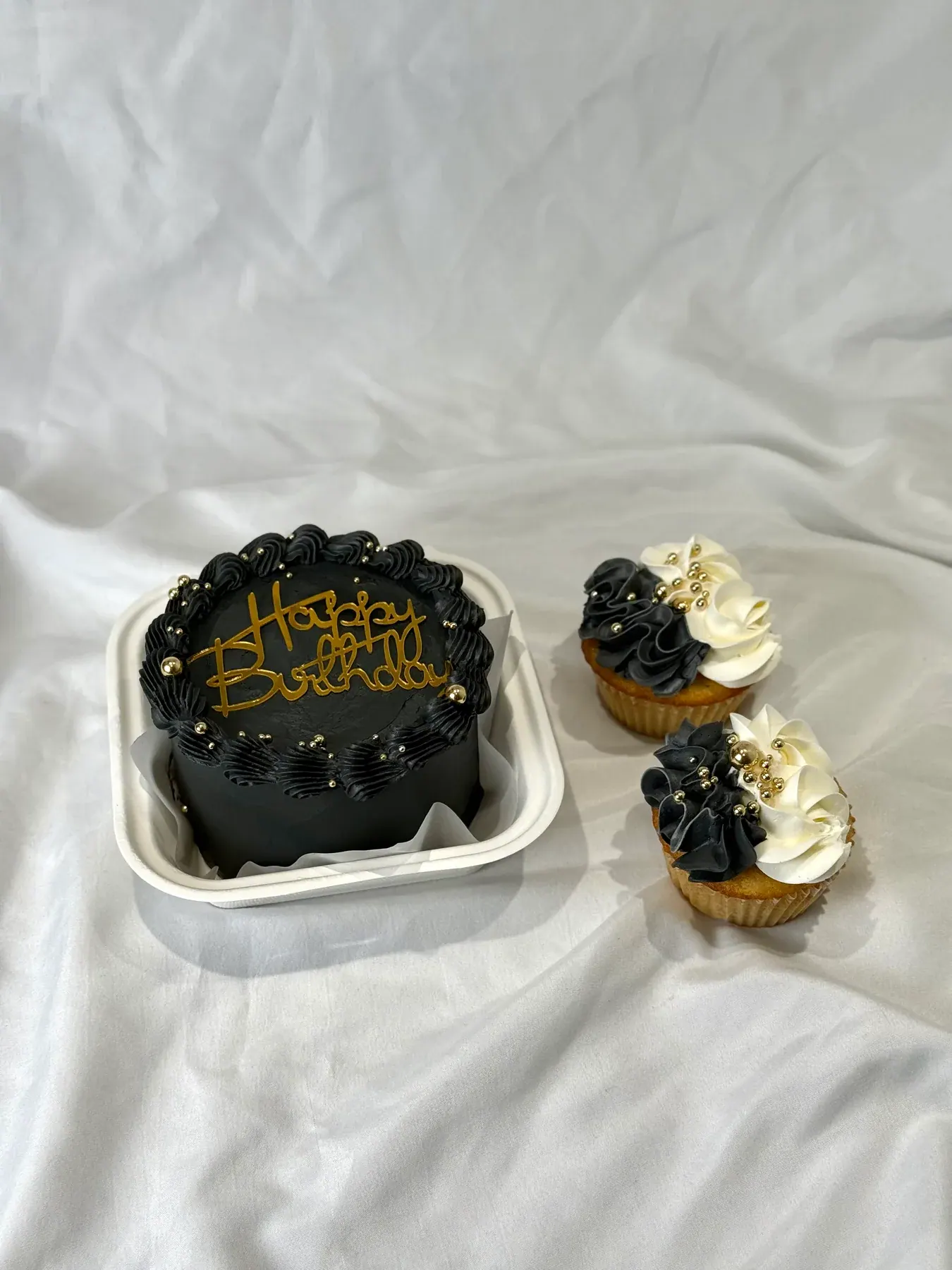 Black bento cake piped with gold Happy Birthday lettering, with two matching cupcakes